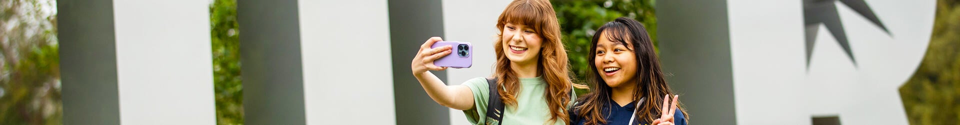 Two female students pose for a selfie in front of the UCR letter sculpture on the UC Riverside campus.