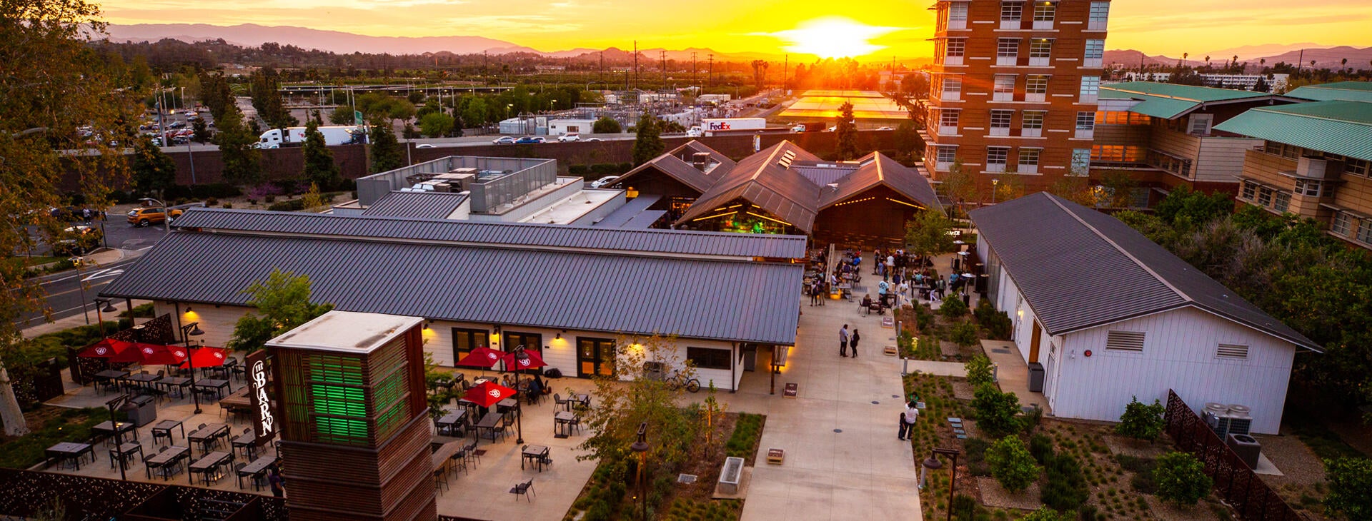 An aerial view at dusk of The Barn on the UC Riverside campus. The sun is setting in the background while people gather and and enjoy the casual bar and grill at UCR which offers comfort food, beer, and live music. 