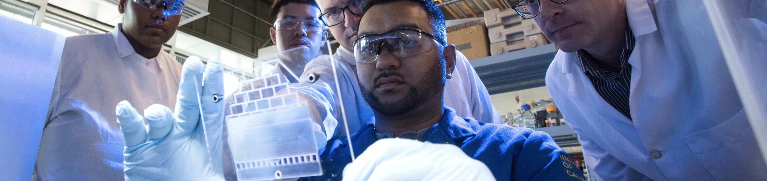 A scientist in gloves and safety glasses holds up a transparent, etched plate while several colleagues look on in a lab setting.