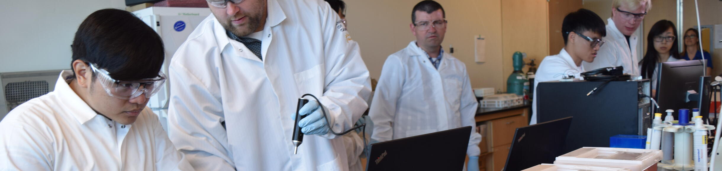 A group of students and instructors wearing lab coats and safety glasses collaborating around laptops and lab equipment in a brightly lit science laboratory.