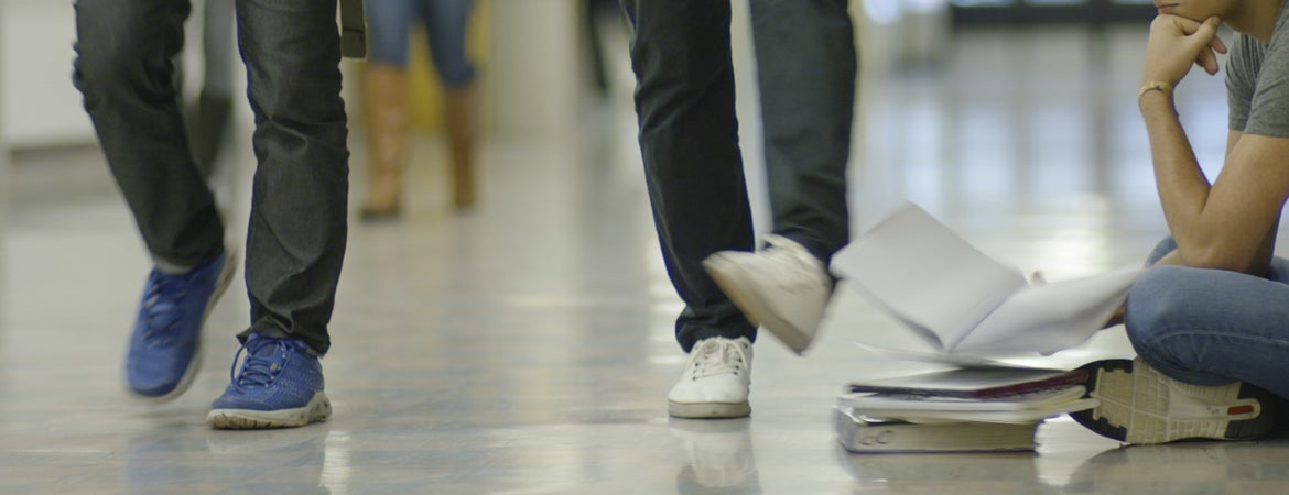 A student kicks the books of another student who is sitting on the floor studying.