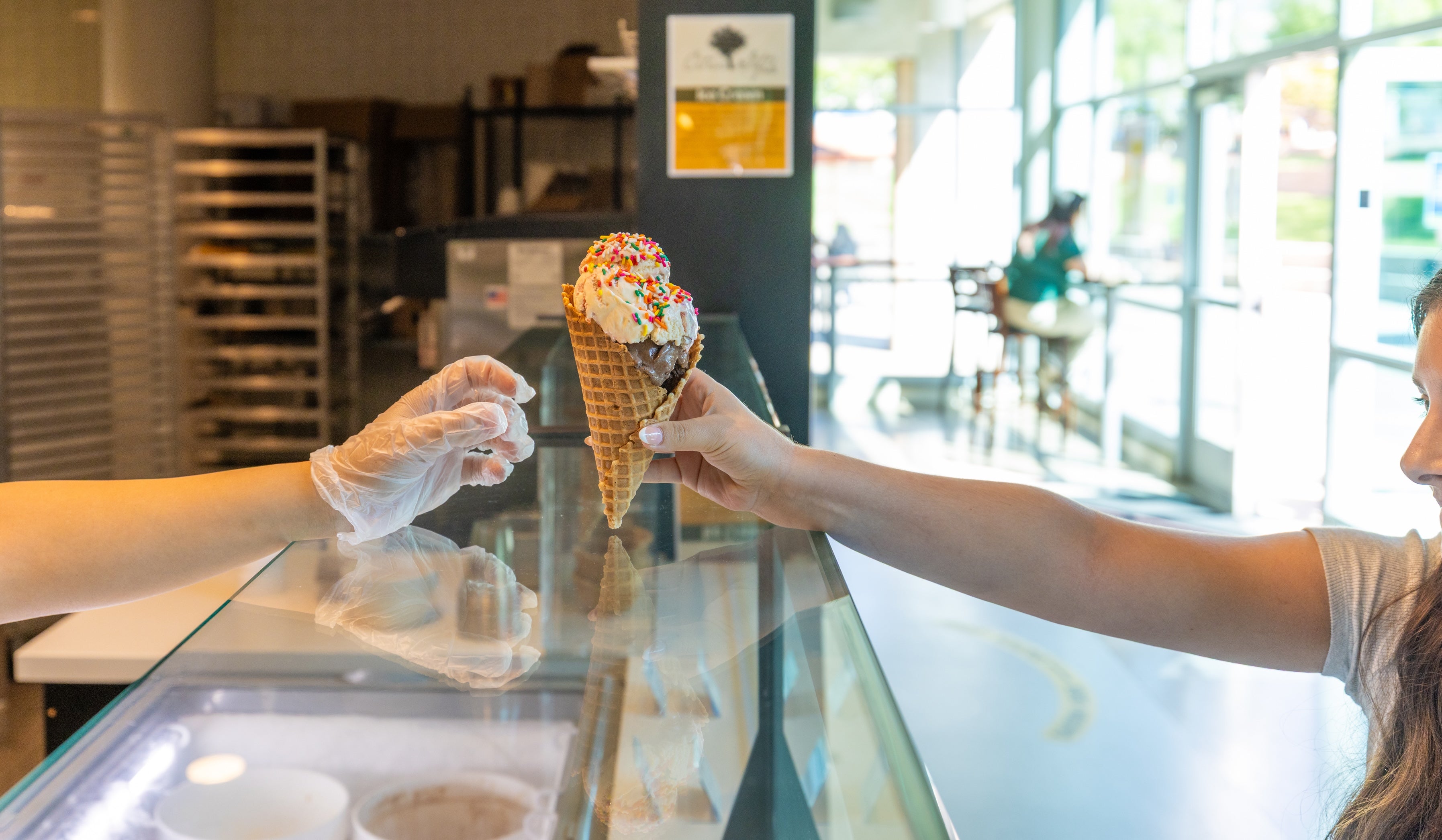A Scoops employee hands a customer a cone with vanilla ice cream and sprinkles.