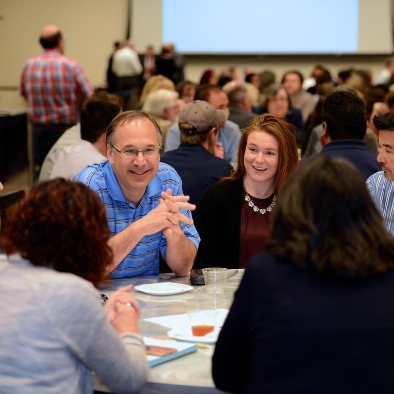 Faculty and staff members talking together at a round table in a room full of poeple sitting together at round tables