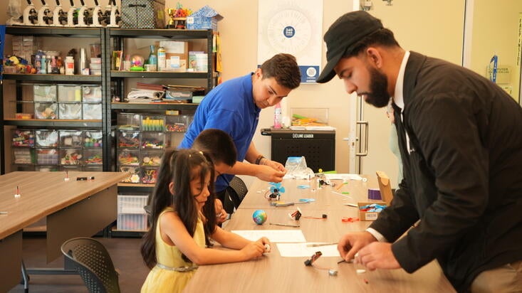 Two adults and two children are engaged in hands-on STEM activities at a table during an Earth Day workshop. They are working with small motors, wires, and craft materials in a classroom with shelves of educational supplies in the background.