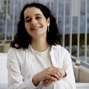 Image of María Abizanda Cardona sitting with both hands on knee. Smiling with a white tshirt underneath a long-sleeved white button down shirt.