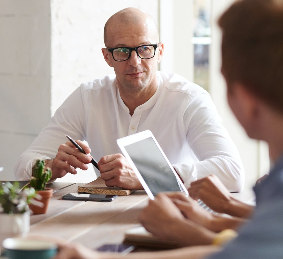 Two White men sitting at a desk with a tablet and and book on the table One man is older with glasses and the other is a side view of a young man