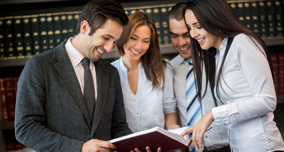 Close up of the faces of two young men and women smiling and looking down at something in a law office with a wall of law books behind them