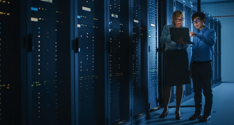 Two IT specialists talking and working on a laptop in front of several server racks