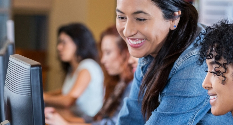 Hispanic female teacher smiling as she shows a highschool age Black girl something on her computer in a class with other students working on computers
