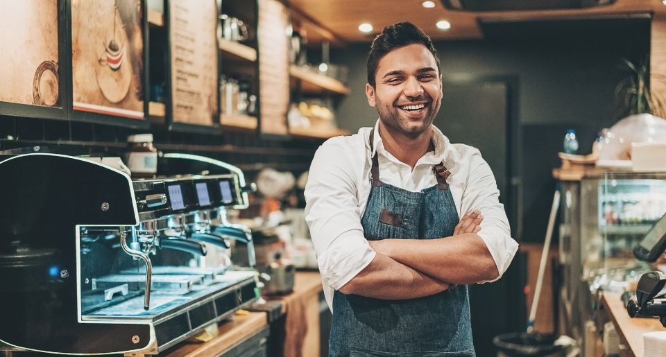 Hispanic male business owner standing in his coffee shop smiling with his arms folded