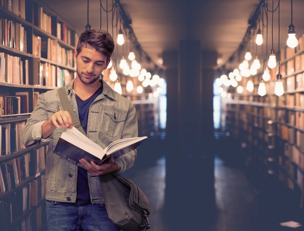 Male college student in library looking at a page of a book that hes holding