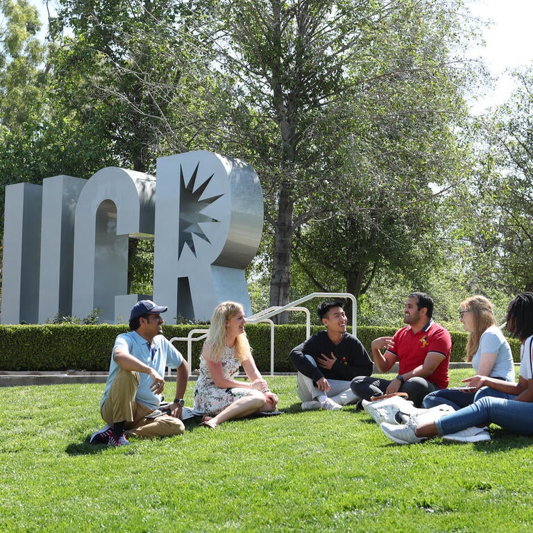 A group of students sit on a grassy area in front of the letter U C and R