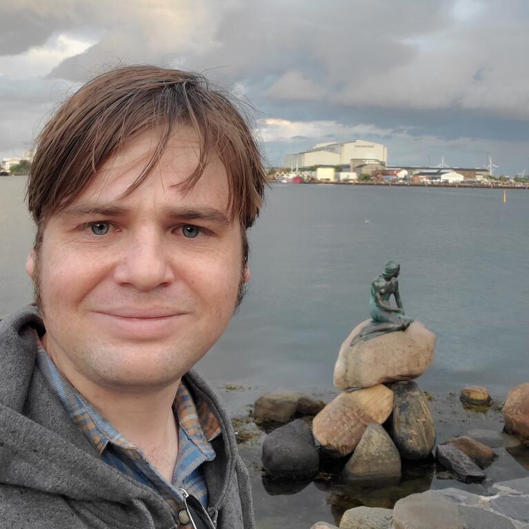 Bryan from the shoulders up, standing in front of the Hans Christian Anderson mermaid statue in Copenhagen harbor.