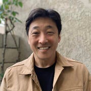 A photo of Jason from the shoulders up, smiling, wearing a black t-shirt and tan jacket