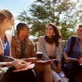 Group of students sitting in sun and chatting with two holding coffee cups