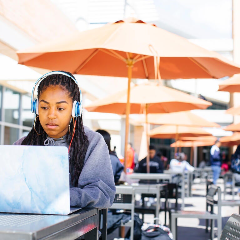 A female student wearing over ear headphones sits at a table while looking at her laptop screen