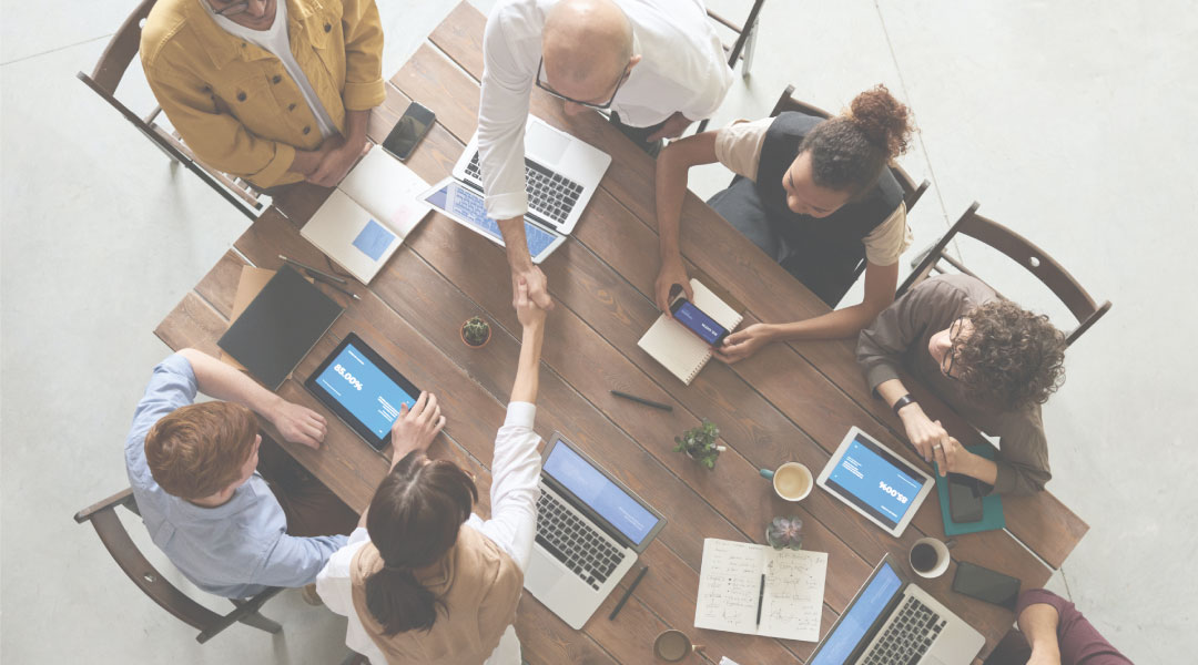 Seven individuals sitting around a table with their laptops, tablets, and notebooks out while two of them- female and male sitting across one another, are shaking hands.