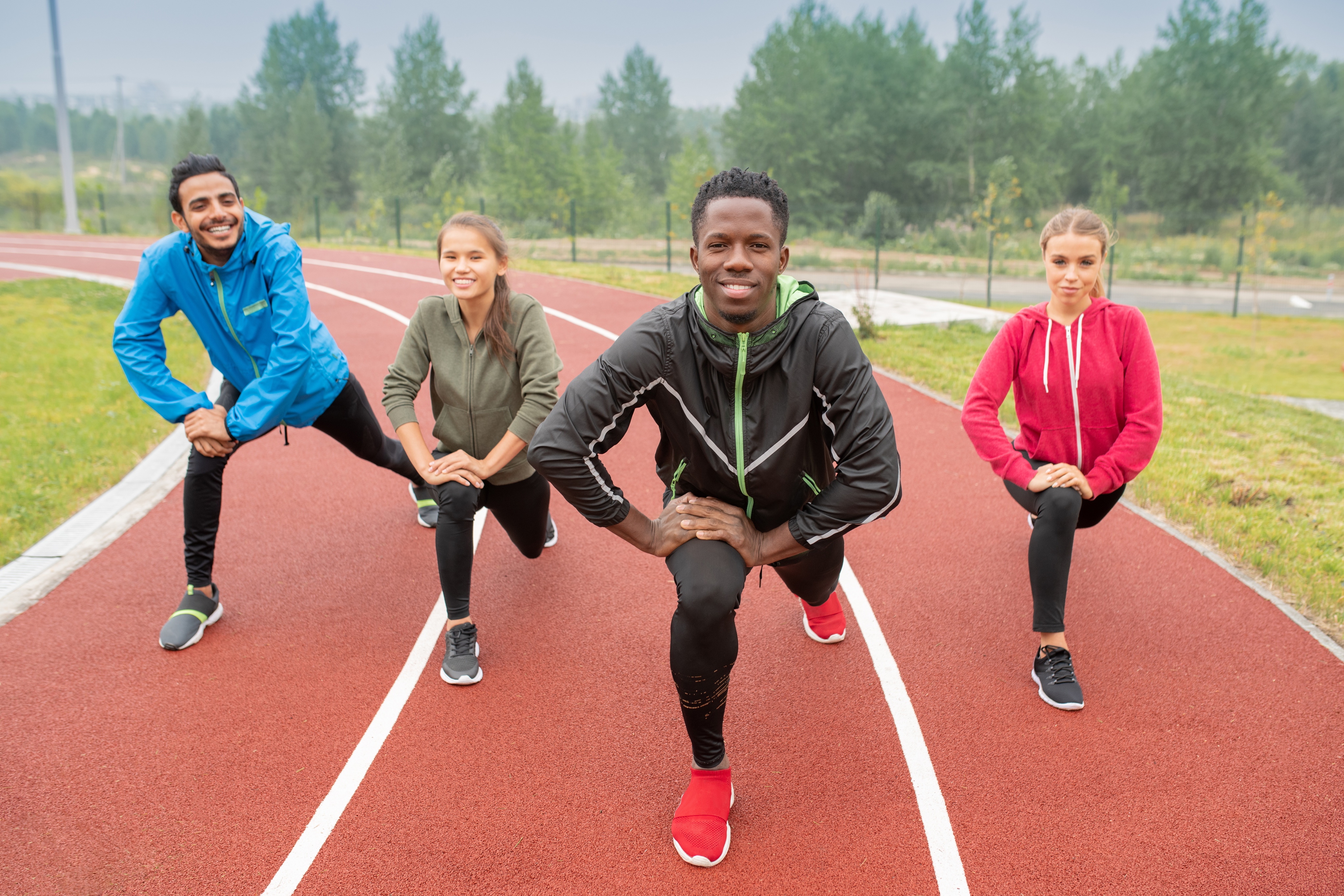 Two men and two women of diverse backgrounds are on an outdoor track doing a knee stretch before running while wearing track suits. before running.