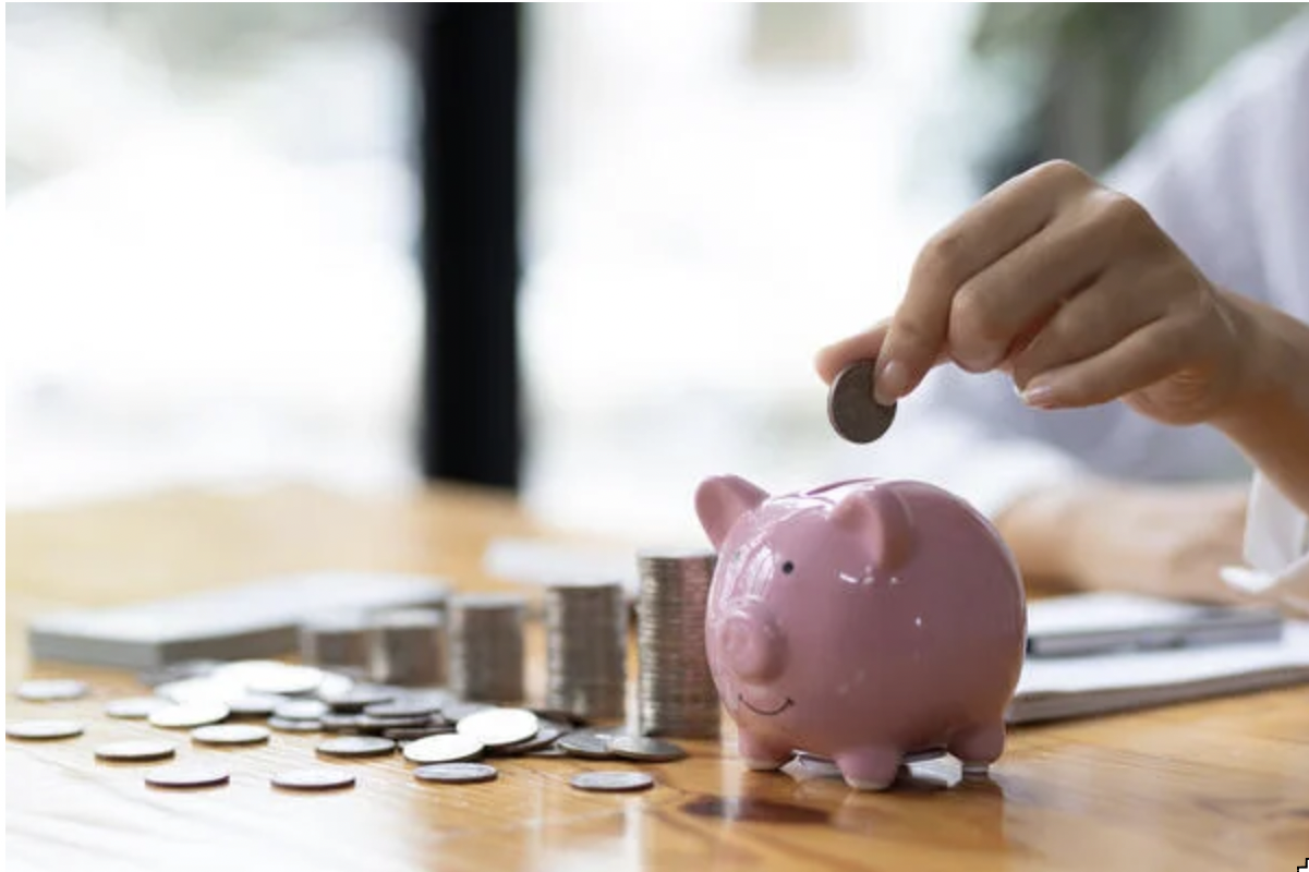 Female hand putting a coin into a pink piggy bank which is on a table next to various size stacks of coins 