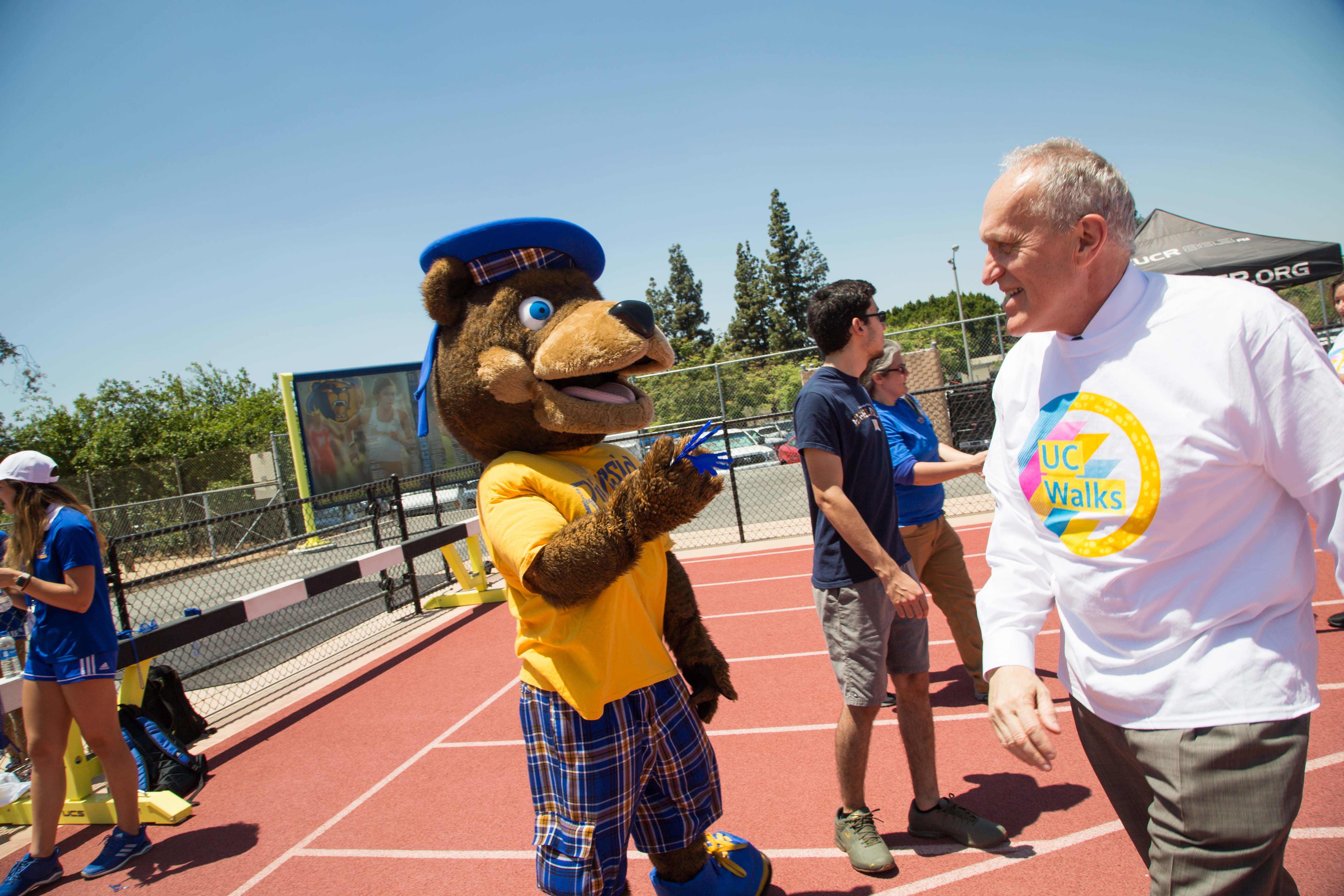 UCR Chancellor Kim Wilcox wearing a white T-shirt with the UC Walks logo being greeted by Scotty the Bear as he walks on the UCR track while participating in the UC Walks event.
