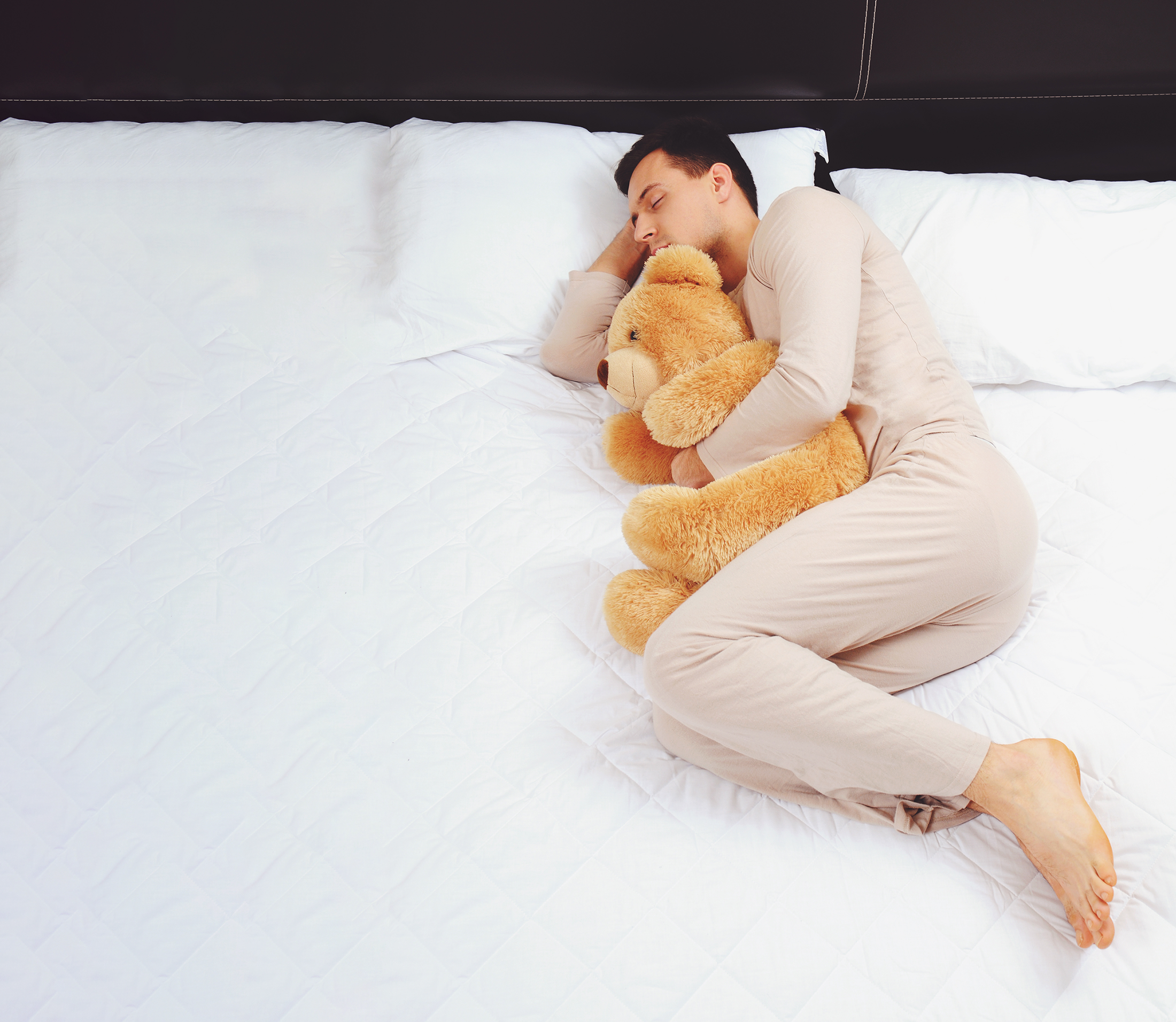 Young man curled up asleep on a bed hugging a fuzzy teddy bear while wearing pink pajamas.