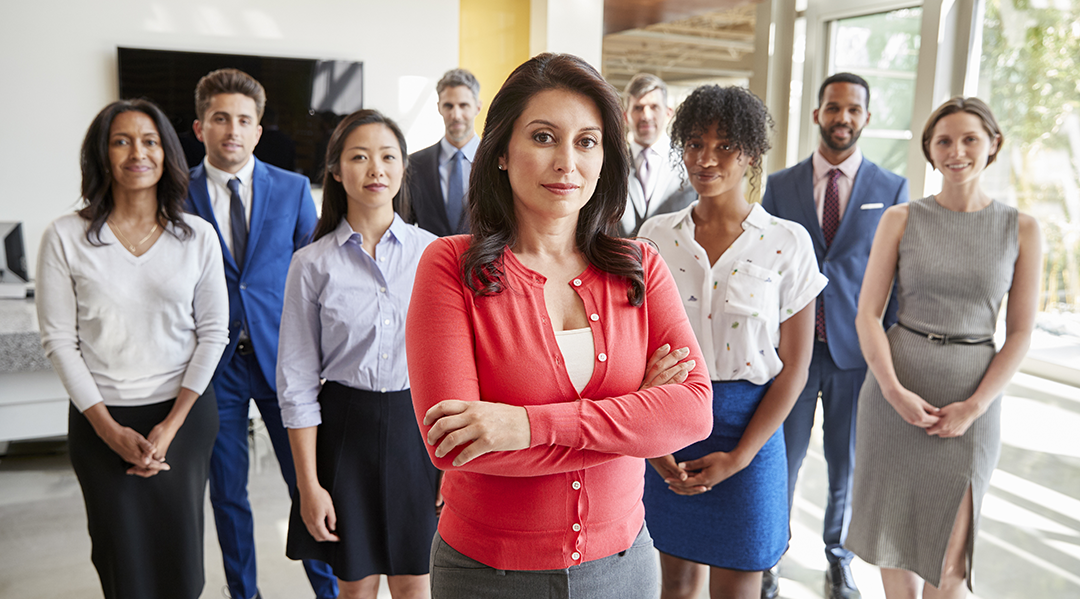 Group of professional people wearing business attire standing together behind their female boss who is standing in the center and crossing her arms