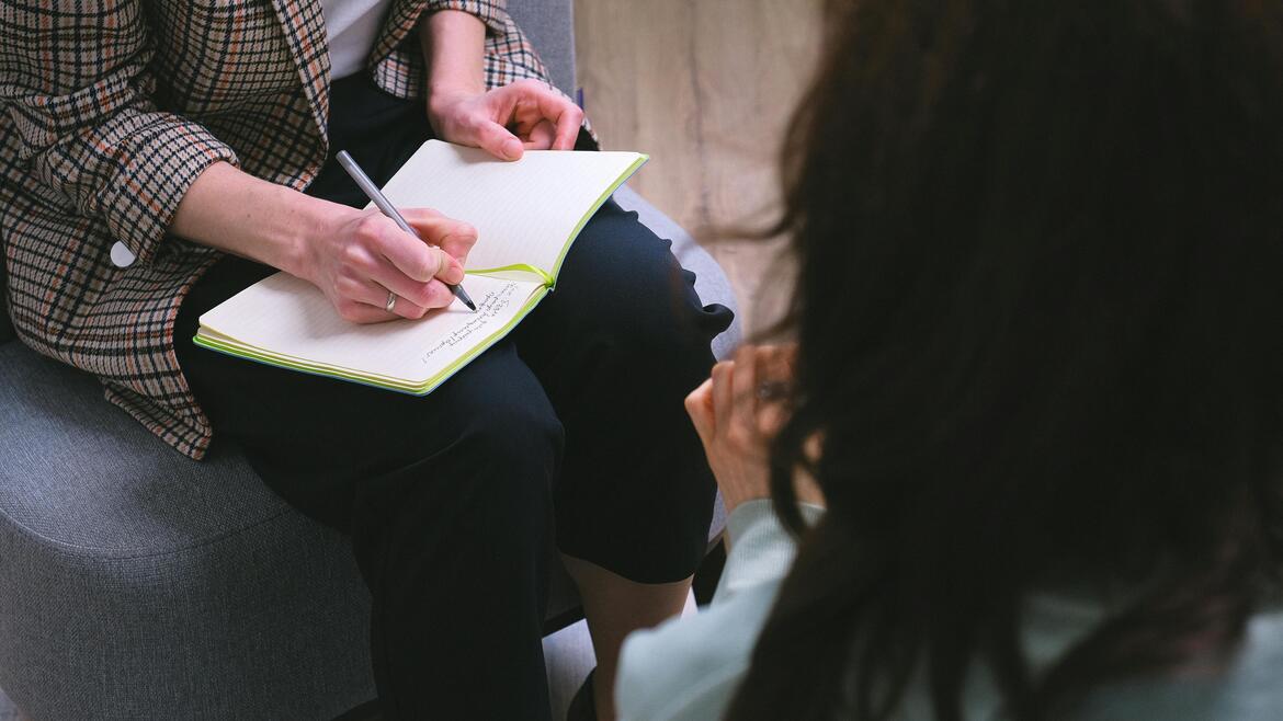 Two women sitting down, one writing on a notebook with a pen, and the other clasping her hands 