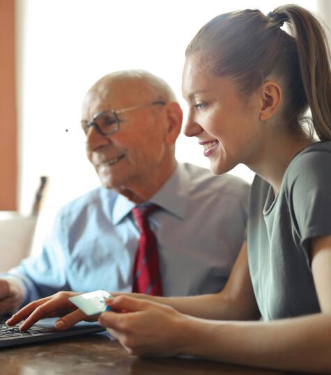 An older man sitting next to a younger woman who is looking at her laptop and seems to be helping him