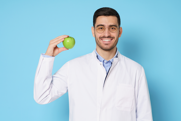 Young smiling doctor with dark hair, beard and glasses is wearing a white smock against a blue backdrop while holding up and apple in his right hand.