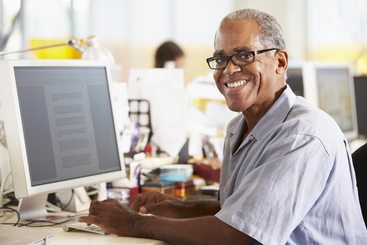 Smiling senior African American man wearing glasses while sitting in front of a computer monitor.
