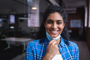 A smiling dark haired woman wearing a blue multi-colored plaid blouse while pulling down a mask from her mouth