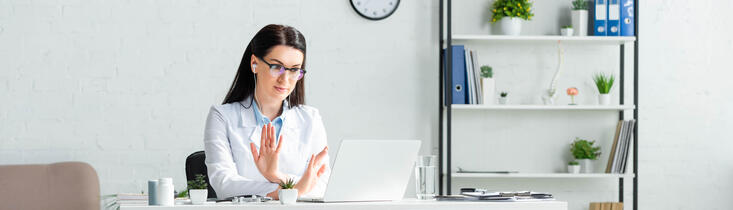 Female health care facilitator wearing glasses and a white coat looking at her computer while making an x with her hands