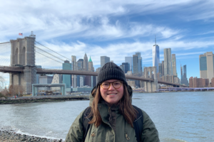 Female, early 20s, with brown hair, wearing a dark green winter coat, a black knitted winter hat, and glasses, standing near water, with the Brooklyn Bridge and the New York City Skyline behind