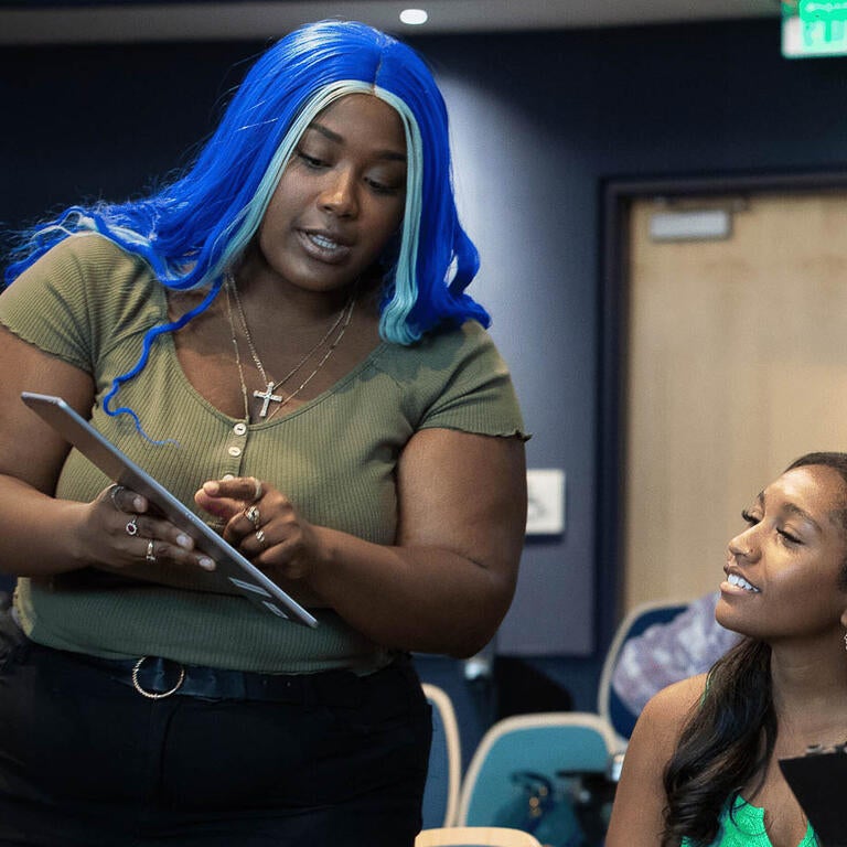 A woman with blue hair holding a tablet and showing screen to three female students