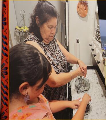 Three images from the Semana de la Mujer exhibit at Tomás Rivera Library: a black-and-white portrait photograph of a young woman from 1958 set against a colorful woven textile border; a color photograph of an older woman teaching a young girl at a kitchen counter; and an illustrated lotería-style card numbered 8, depicting a crowned woman holding a baby, titled 'La Hermana Mayor'.