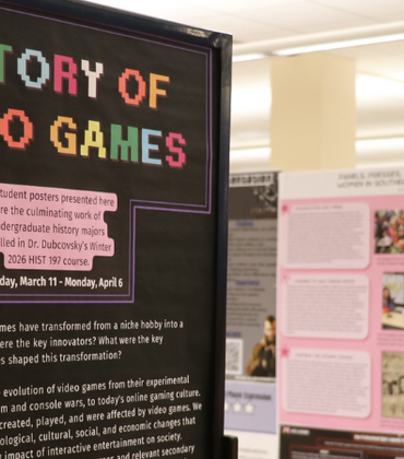 This exhibition, titled "History of Video Games," showcases student research posters from Dr. Dubcovsky’s Winter 2026 HIST 197 course. The display features a prominent pixel-art sign explaining the course's focus on the cultural evolution of gaming, with the exhibition running from March 11 to April 6.
