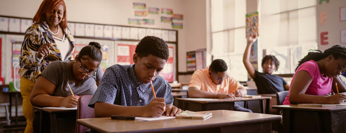 A teachers walks among students doing work at their desks.
