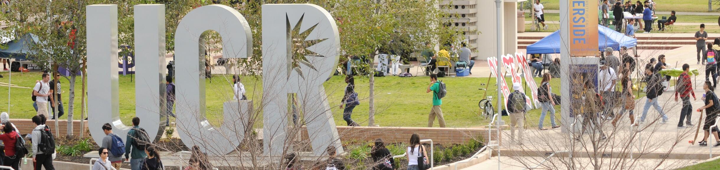 Large "UCR" letters on a green campus lawn with a tall, with the Bell Tower in the background, surrounded by students walking.