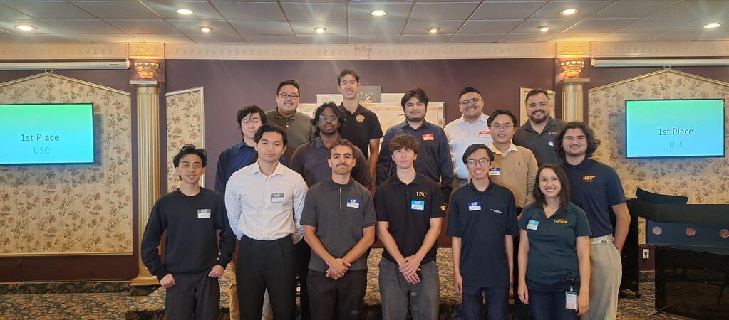 Group photo of student teams from multiple universities standing together indoors after a competition. Two screens in the background display '1st Place USC'. Participants are wearing name tags and a mix of university-branded shirts and business casual attire.