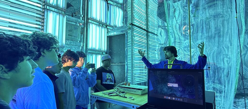 A group of five high school students on a STEPCon (Science and Technology Education Partnership Conference) tour are gathered in the world's largest indoor atmospheric processes chamber at the UC Riverside CE-CERT Atmospheric Processes Laboratory. The students are intently focused on a researcher in a blue jacket, who is enthusiastically explaining a concept with his arms raised, standing near a laptop. The massive chamber is dramatically lit by arrays of intense cyan/blue UV lights