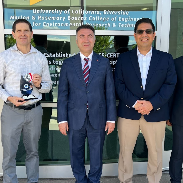 A photo of five men standing in a line outside of the CE-CERT building at UC Riverside. From left to right: Arun Raju, Don Collins (holding a plaque), Prof. İdris Demir, Alfredo Martinez-Morales, and Musa Yilmaz. The photo was taken during Prof. Demir's visit to explore opportunities for international collaboration in energy research and academic exchange.