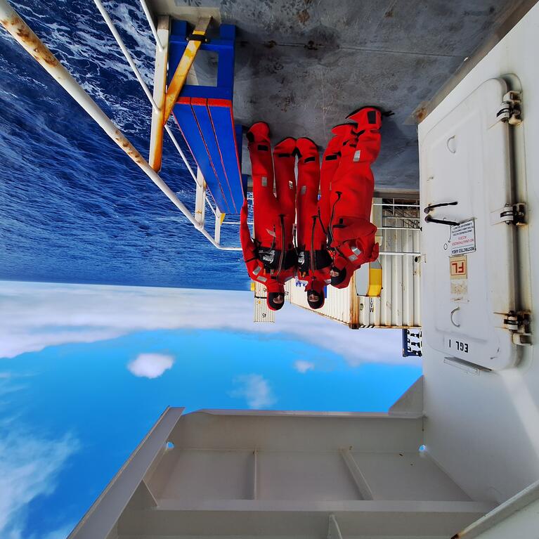 Three researchers wearing bright red immersion suits stand on the deck of a ship, with the open ocean and partly cloudy blue sky in the background. The group is positioned near a white metal door and railing, with containers and equipment visible on the deck.