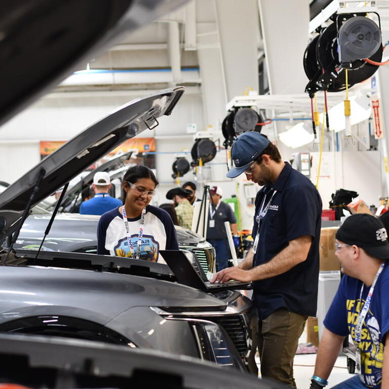 Four UCR EcoCAR team members work together in a garage during the EcoCAR EV Challenge. One student types on a laptop placed on the engine bay of a Cadillac LYRIQ with the hood open, while another smiles and observes. Two others stand nearby, one seated and one wearing safety gloves, as additional teams work in the background of the well-lit facility.
