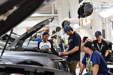 Four UCR EcoCAR team members work together in a garage during the EcoCAR EV Challenge. One student types on a laptop placed on the engine bay of a Cadillac LYRIQ with the hood open, while another smiles and observes. Two others stand nearby, one seated and one wearing safety gloves, as additional teams work in the background of the well-lit facility.