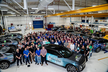 A large group of students and mentors from all participating teams pose together inside a spacious auto workshop, standing around multiple Cadillac LYRIQ vehicles, including the EcoCAR EV Challenge car in the center foreground.