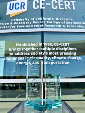A glass award displayed in front of the University of California, Riverside's CE-CERT building, reflecting the text "University of California, Riverside Bourns College of Engineering Center for Environmental Research & Technology." The award is from the South Coast Air Quality Management District, recognizing the center for "Leadership in Air Quality." The background text highlights CE-CERT's mission to address air quality, climate change, energy, and transportation challenges