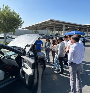 The UCR EcoCAR team presentation during CE-CERT's Open House. A group of approximately 10 high school or university students are gathered outdoors on a sunny day in a parking lot. They are listening to a man in a blue shirt standing over the open hood of a dark car, which has its driver's side door open. The EcoCAR project information is visible on the hood. A large solar panel structure is visible in the background.