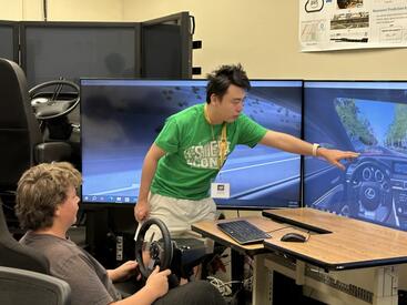 A student sits in the driver's seat of the Transportation Systems Research (TSR) driving simulator, holding a steering wheel, while a researcher in a green shirt points at a large monitor displaying a driving simulation. The setup features multiple large screens surrounding the driver to create an immersive experience.