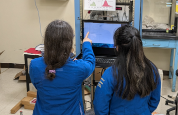 Two women in blue lab coats stand in front of a computer screen displaying a graph. One of them points to the screen as they discuss the data.