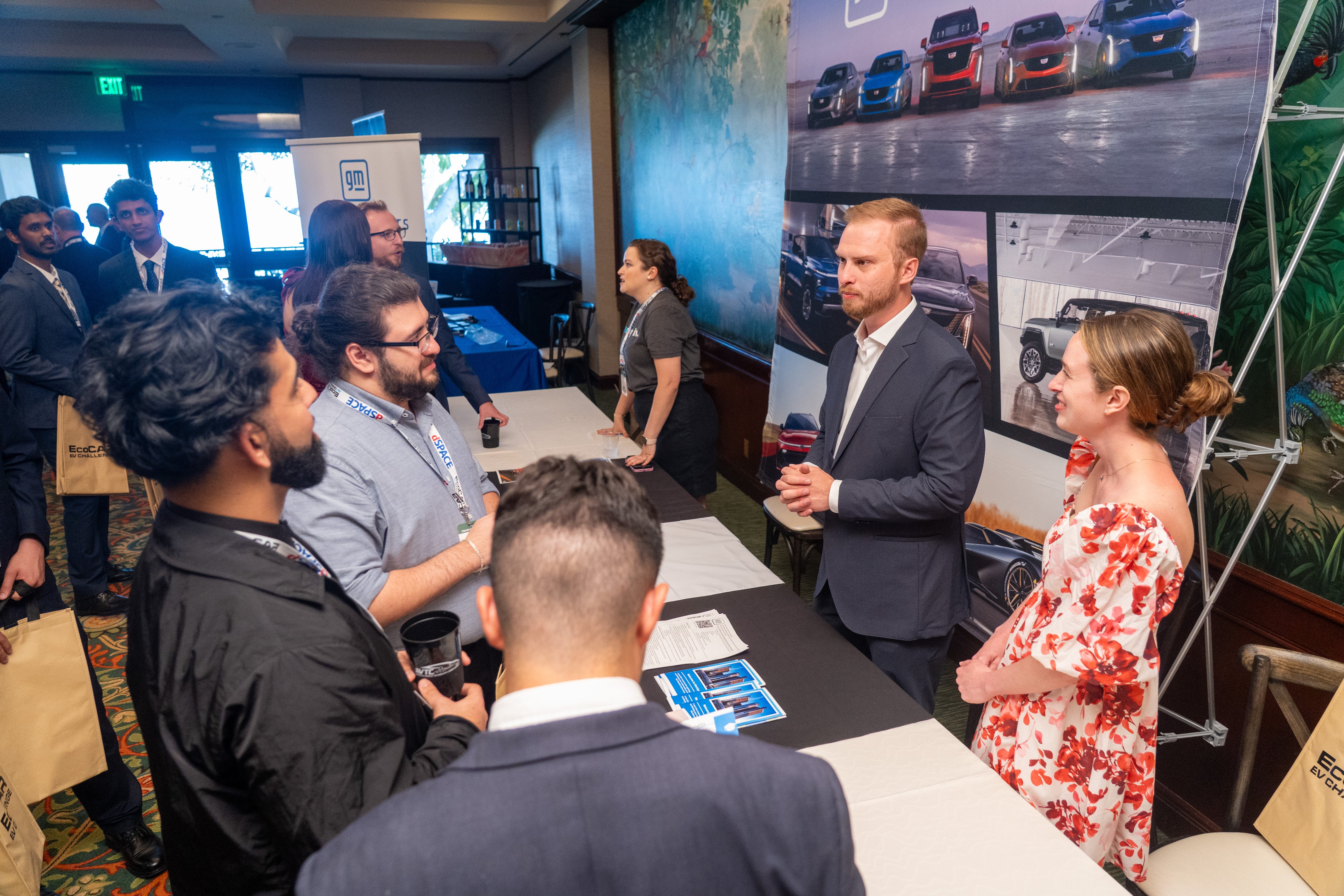 UC Riverside EcoCAR EV Challenge team members engage in conversation with representatives at a career fair booth featuring General Motors branding and automotive displays. Team members hold event tote bags and promotional materials while networking with industry professionals.